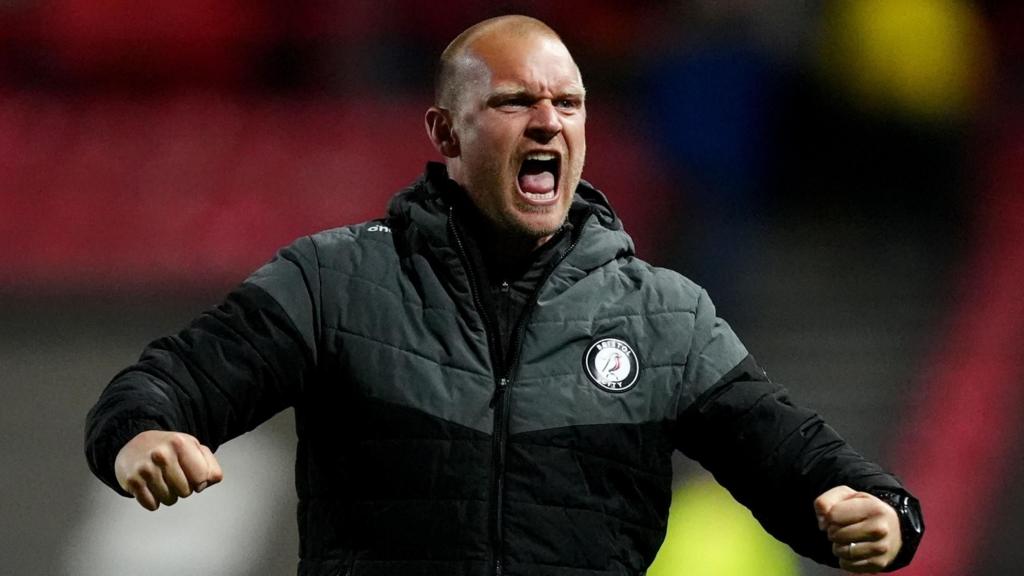 Liam Manning with his arms bent outwards at chest level. He is shouting and has clenched fists. He is wearing a Bristol City puffer jacket.
