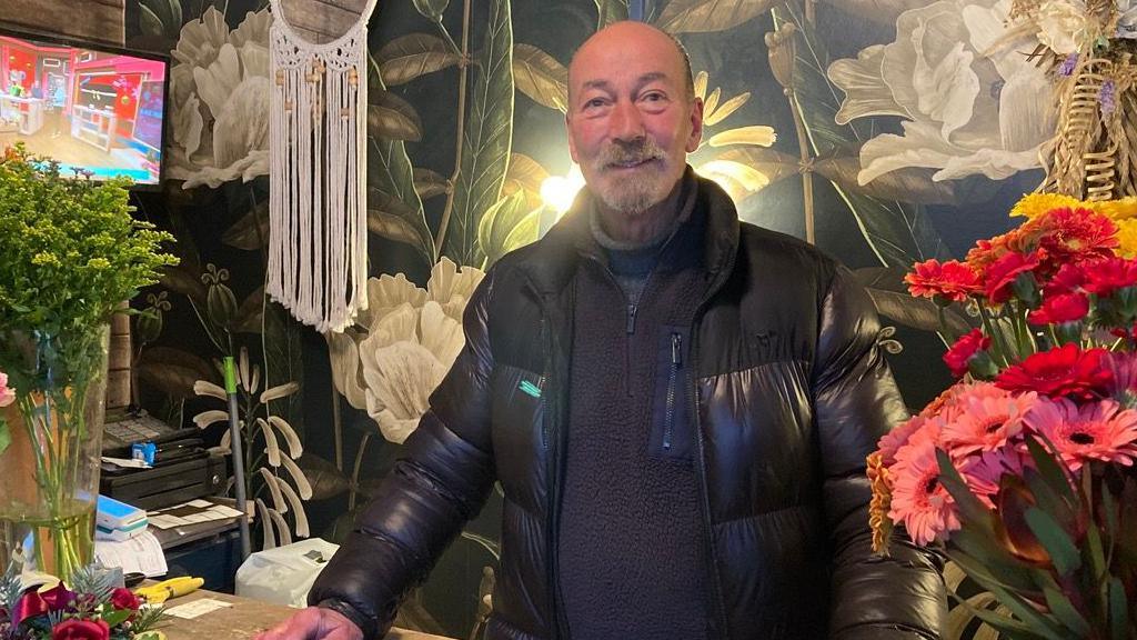 Richard Sharman is wearing a black jumper and a black puffer jacket. He is standing next to bunches of flowers with floral wallpaper behind him