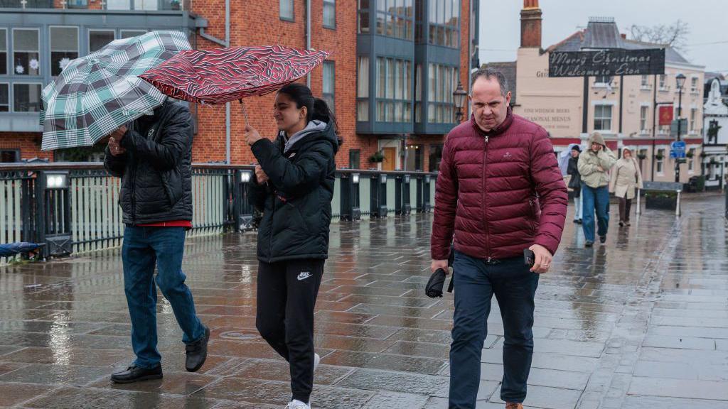 A woman struggles with an umbrella as a man walks on wet ground in Windsor 