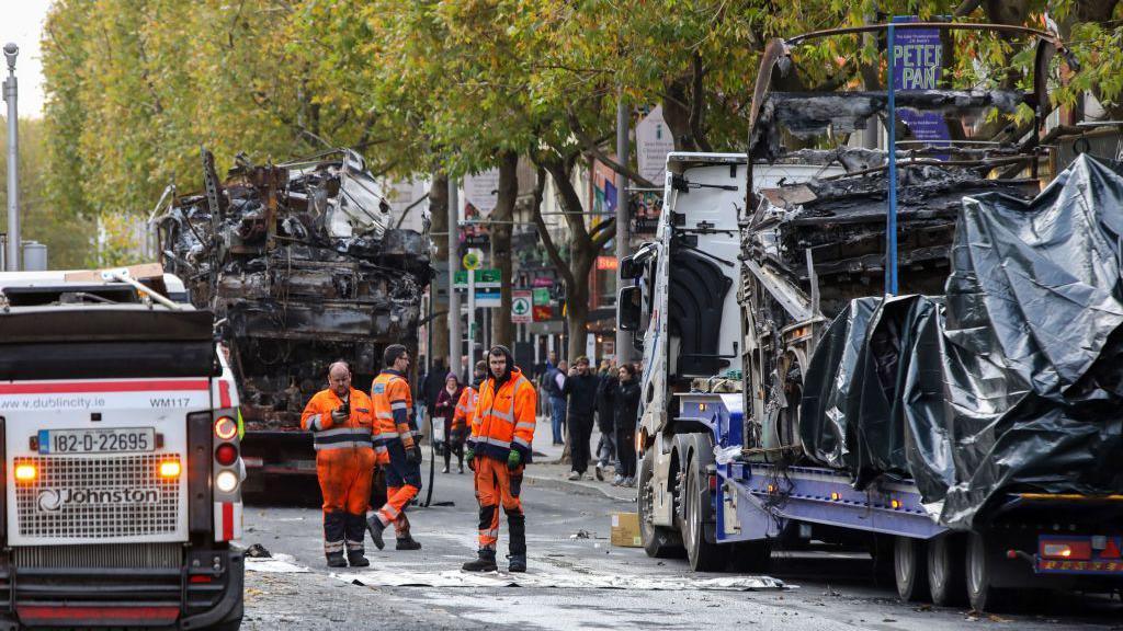 Workers clear debris from the road as the shells of burnt out buses wait to be removed from O'Connell Street