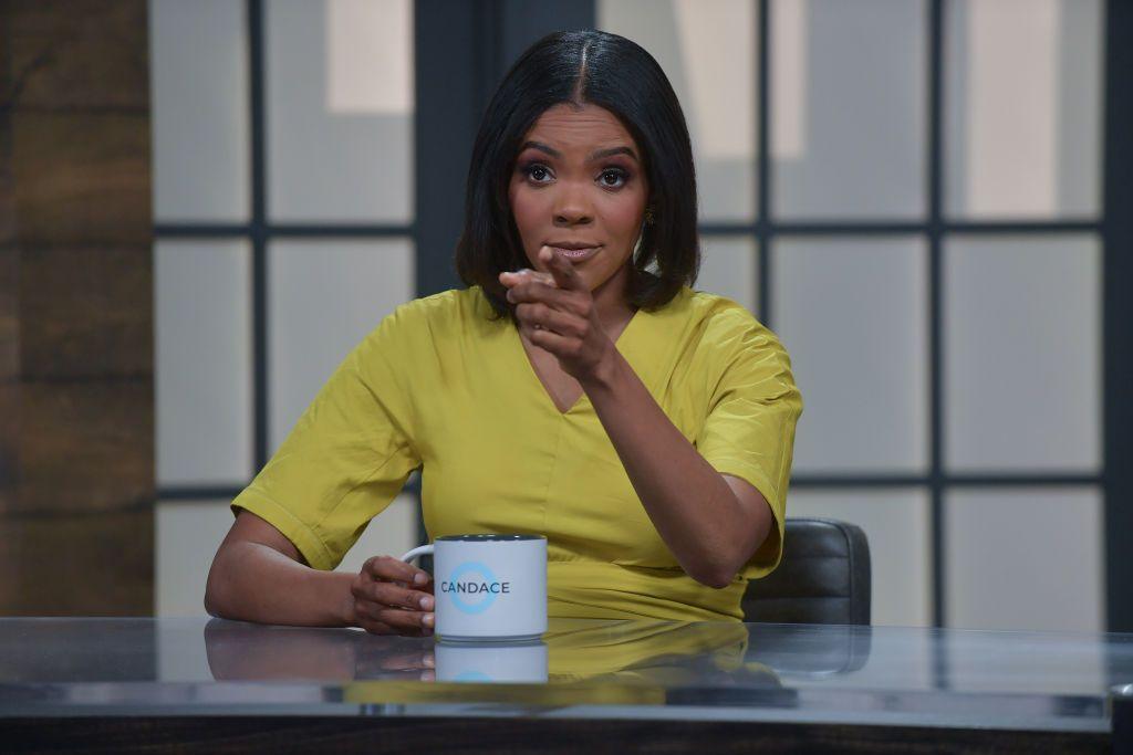 Owens behind a desk holding a mug which says "Candace" on it, pointing towards the camera.
