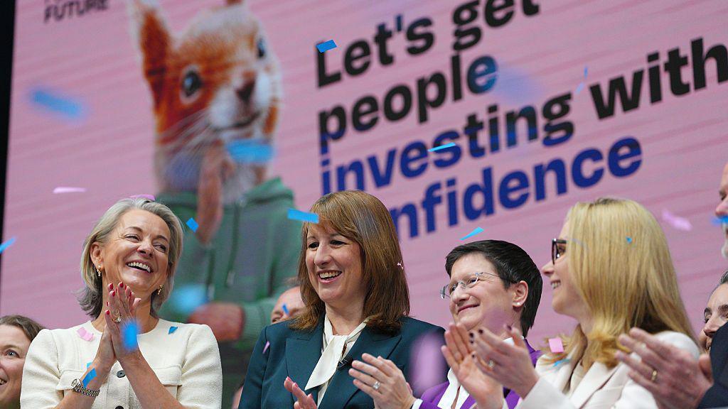 Chancellor Rachel Reeves, Julia Hoggett, CEO of London Stock Exchange, and Lucy Rigby, Economic Secretary to the Treasury, are seen at the launch the investment campaign, at the London Stock Exchange on April 23, 2026. They are clapping at the opening of the market with the advert on a large screen behind them.
