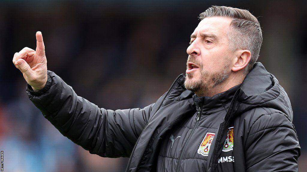 Jon Brady with short black hair and beard pointing on a football pitch