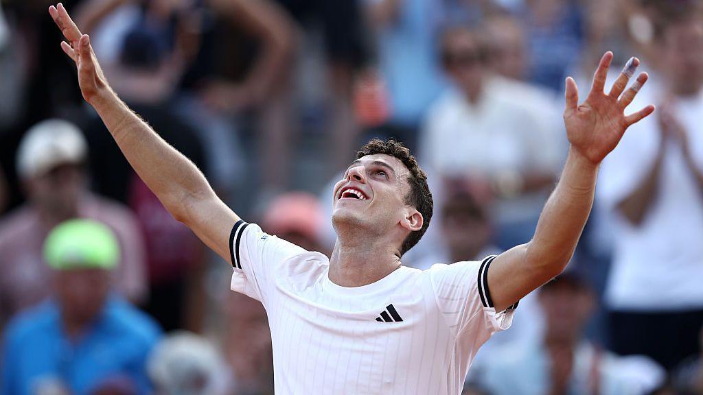 Juan Manuel Cerundolo, wearing a white t-shirt, raises his arms in the air and looks to the sky, smiling, in celebration