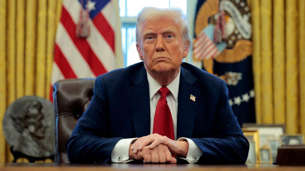 Donald Trump sits at a desk wearing a dark suit with a red tie and with his hands folded in front of him. Behind him is an American flag.