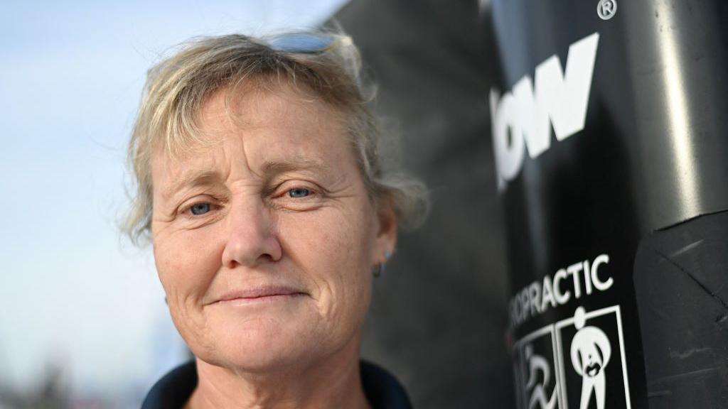 Pip Hare poses for a photograph aboard her Imoca monohull "Medallia" at the starting site of the 10th edition of the Vendee Globe around the world monohull solo sailing race, at the Vendee Globe village in Les Sables-d'Olonne, western France. Her blonde/grey hair is tied back and she is wearing a navy jumper.