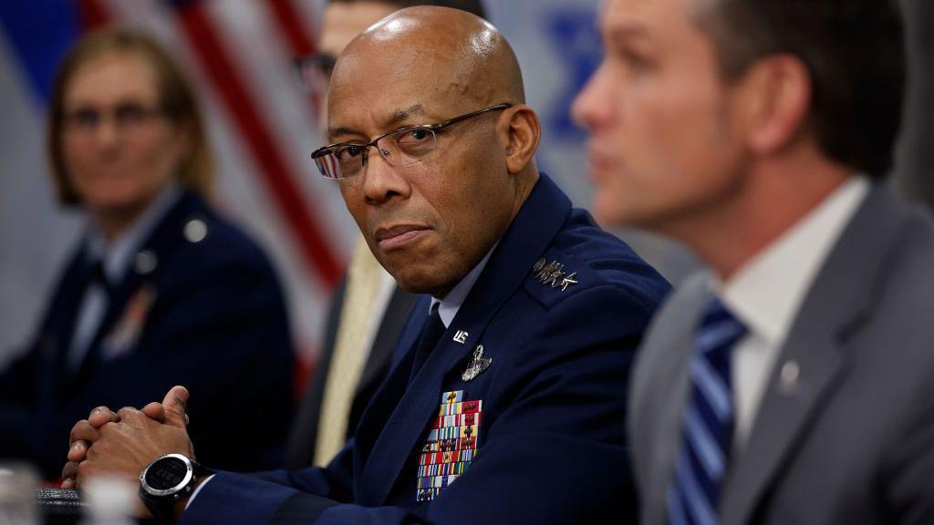 CQ Brown in navy blue uniform and glasses, with hands folded looks over at Pete Hegseth in grey suit