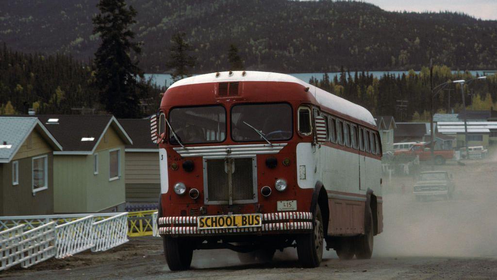 A red school bus driving on a residential road with a lake and forest in the background, photographed in Saskatchewan's Uranium City in 1975. 