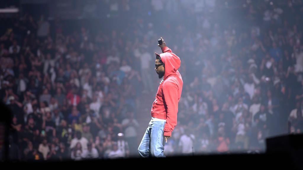 Kendrick Lamar performs on stage in jeans and a red hoodie