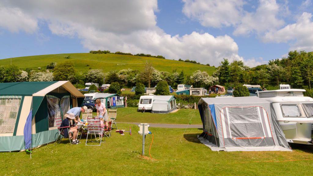 Three people sitting round a small table next to a large tent, with a campsite in the background