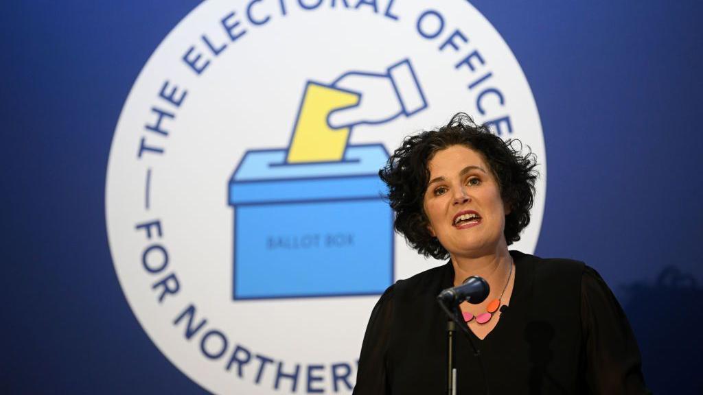 Claire Hanna speaking at a microphone in front of the logo of the Electoral Office for Northern Ireland