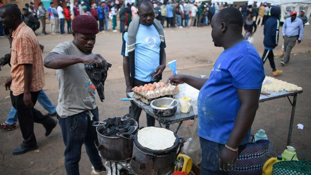 Three men stand near a charcoal heater at a vendor's stall. The vendor is preparing chapattis.