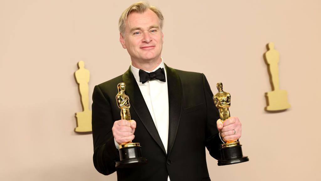 A smiling man wearing a tuxedo and holding his two Oscars in his hands.