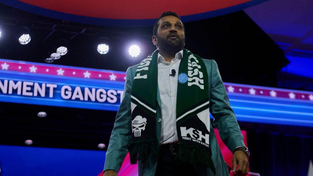 Kash Patel wears a green blazer and green scarf with his name on it. It also has a skull with hair similar to Donald Trump's on it