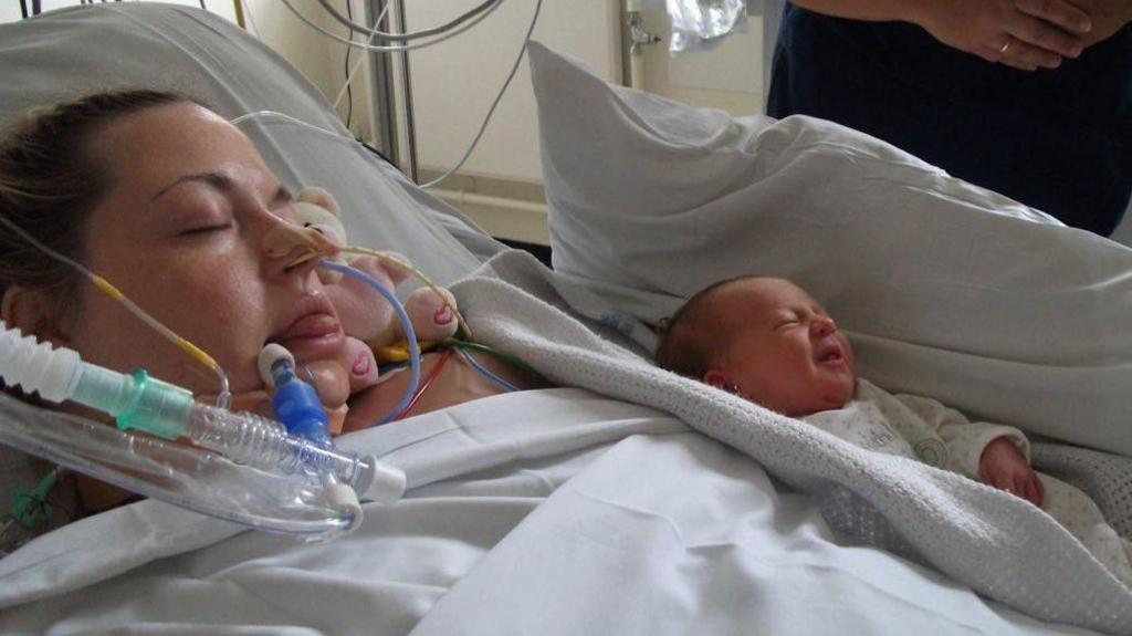 A woman in a hospital bed with the white sheet pulled up to her chin. Her hair, which is brown, is pulled back from her face. She is not conscious and there are tubes entered into her nostrils, and by her mouth. A newborn baby, with a small amount of dark hair and wearing a white babygrow, is lying next to her with a pillow along side and with a white blanket around it. A person is visible standing to the right of the frame, wearing dark blue, but only part of their mid-section and hands can be seen.