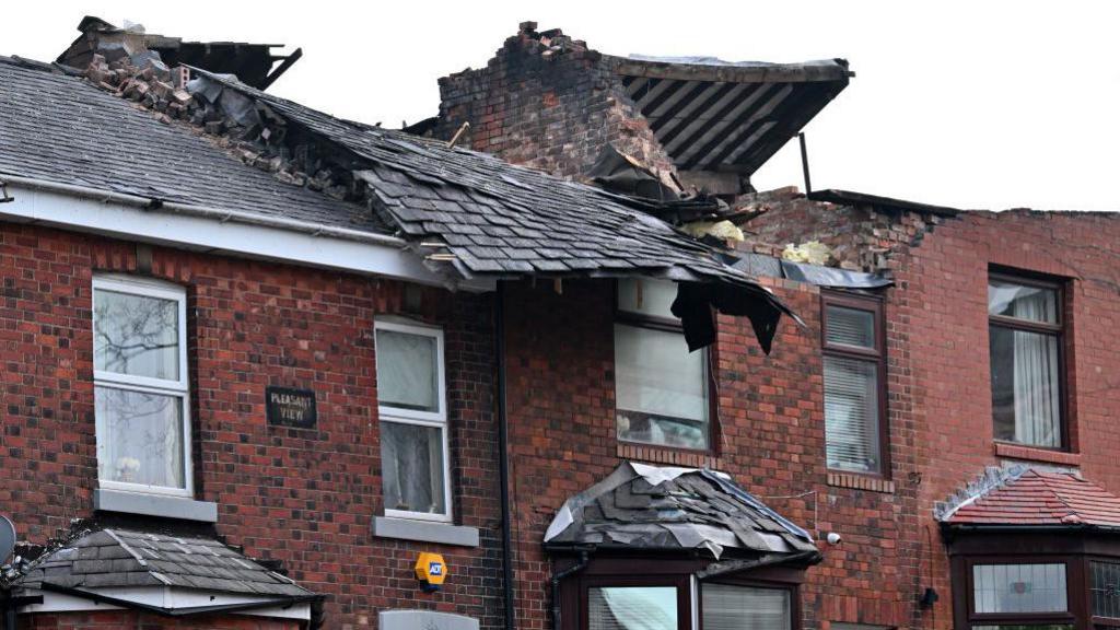 A row of red-brick terraced houses with tiled roofs which have been badly damaged in a storm. 