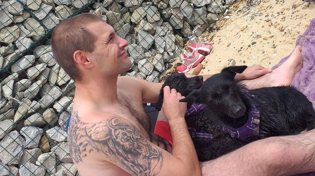 A topless man sits on the beach with two black puppies wearing purple collars on his lap