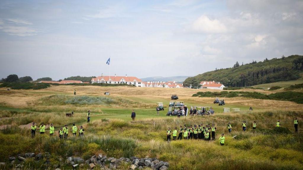 A photo of Turnberry from 2018 when Trump visited as president. In the distance you can see the red roof of the famous hotel. In the middle distance are golf buggies. We assume Trump is in one of them. In the foreground dunes are dozens of police wearing yellow jackets.