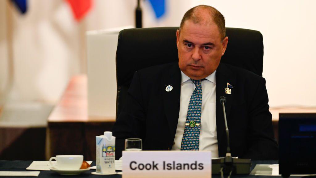 Mark Brown sits in a chair looking serious. In front of him on a table are some cups, a microphone, and a tag that reads "Cook Islands".