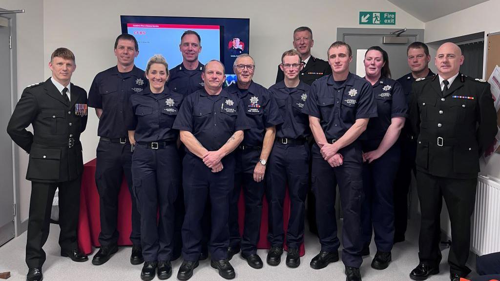 A dozen people in fire service and staff uniforms stand to pose for a photo in the refurbished fire station. 