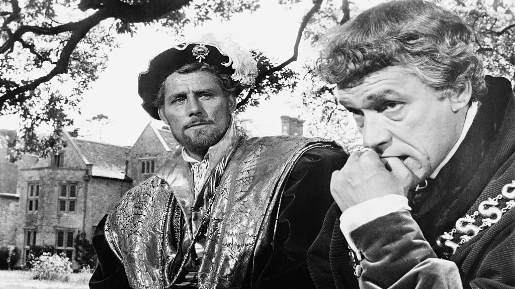 In black and white, Robert Shaw is dressed in a very elaborate regal costume with felt hat and feather. He is looking quizically at Paul Scofield, dressed less elaborately as befits Sir Thomas More's character. The latter is biting his nails. They sit in a garden by a very beautiful medieval stone house. There are trees.
