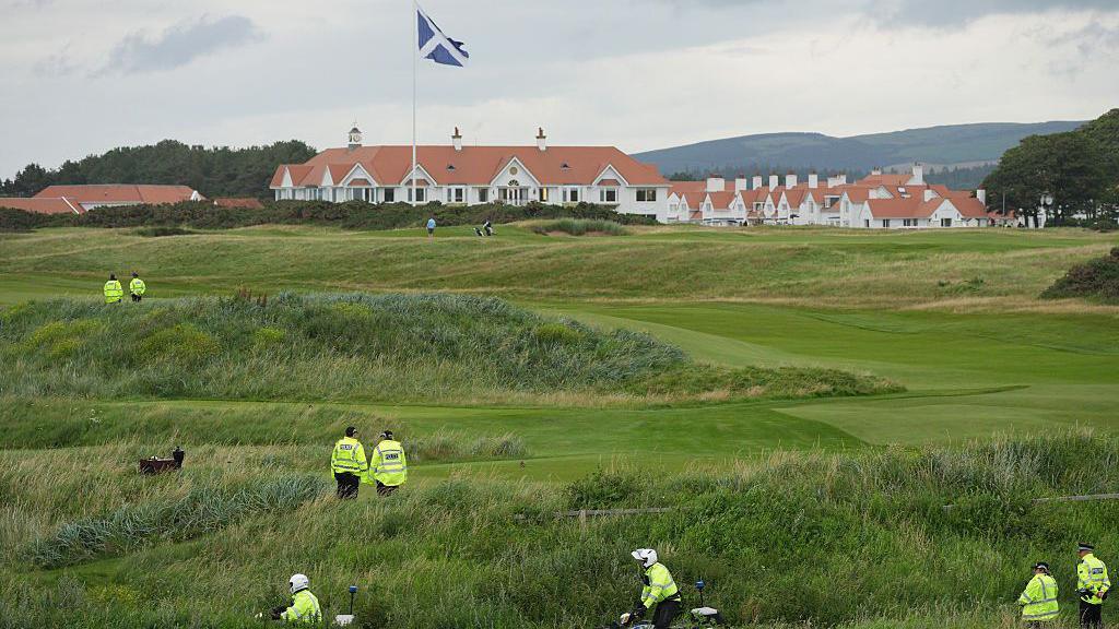 Police officers, visible by hi vis jackets spotted across a golf course. In the background is a white building with a sandstone red roof behind a saltire.