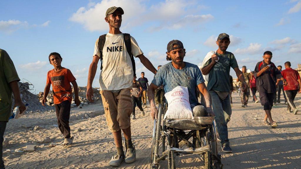A man walks alongside another man in a wheelchair, who is carrying food aid in a white bag on his lap