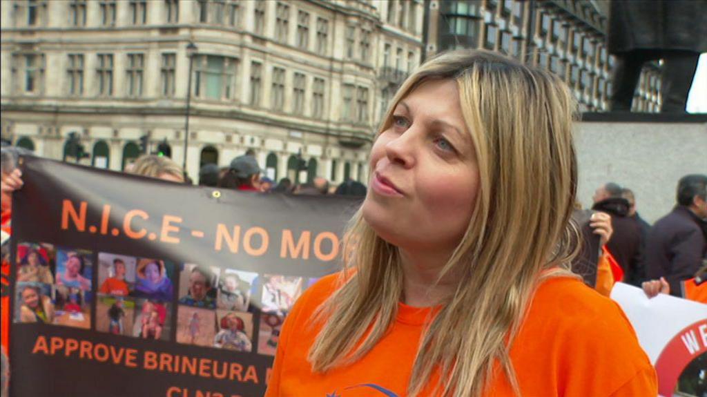 A woman with blonde hair and wearing an orange T-shirt stands outside. Demonstrators can be seen in the background, holding a banner with 