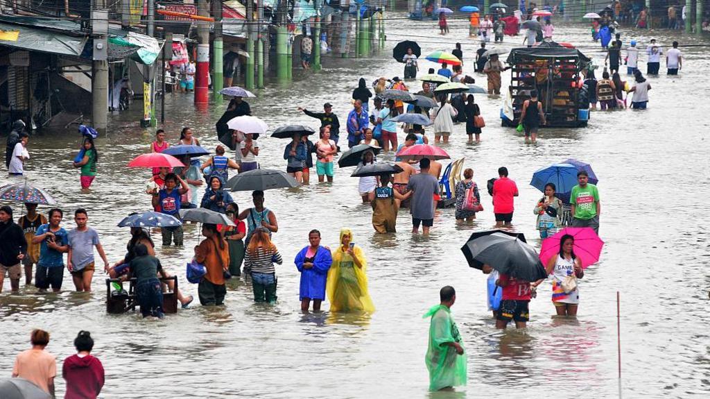 Dozens of people holding umbrellas and wearing raincoats wade through a heavily flooded Manila street
