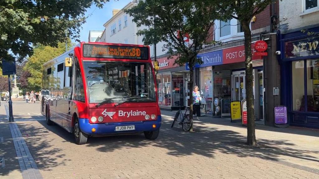 A red and blue, single-decker Brylaine bus runs through a narrow pedestrianised street in Boston. To the right are shops including WH Smith and a Post Office