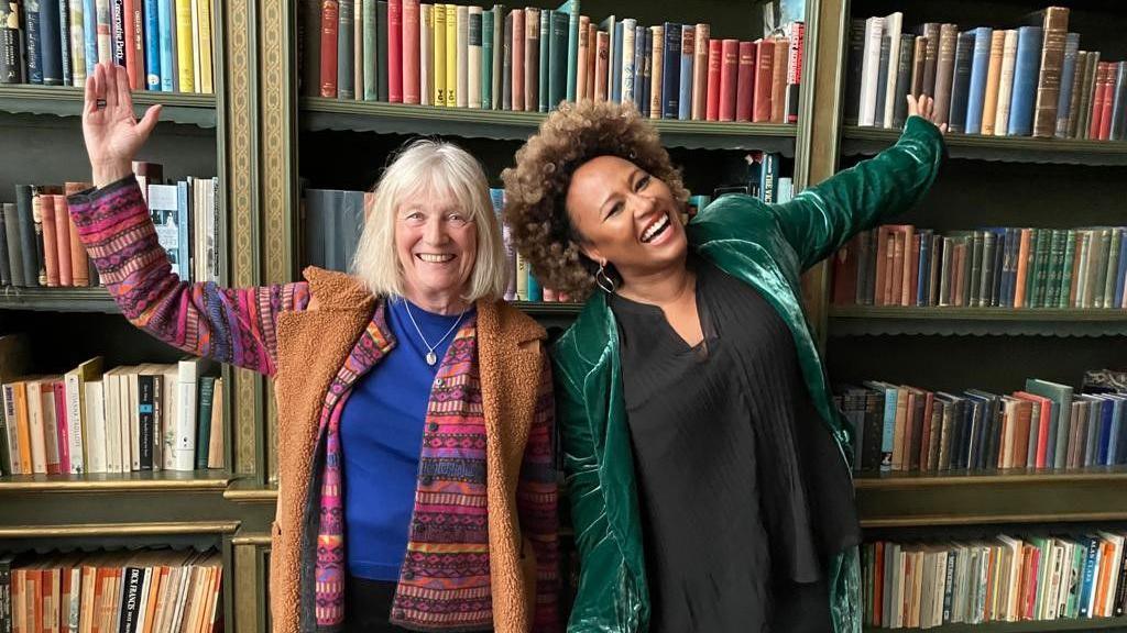 A woman with grey hair in a multicoloured top and brown furry cardigan and a woman with brown curly hair a black top and green jacket standing in front of bookshelves filled with books with their arms stretched wide