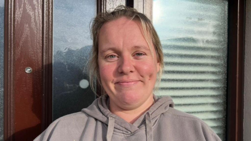 A woman with blond hair stands smiling in front of a house's front door.She is wearing a grey hooded top