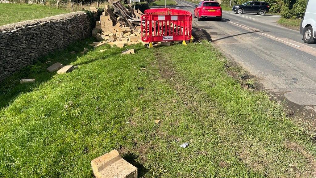 Indentations in the grass verge and scattered debris next to the collapsed bus shelter on Cirencester Road. Red barriers surround the rubble and a red car is being driven on the road