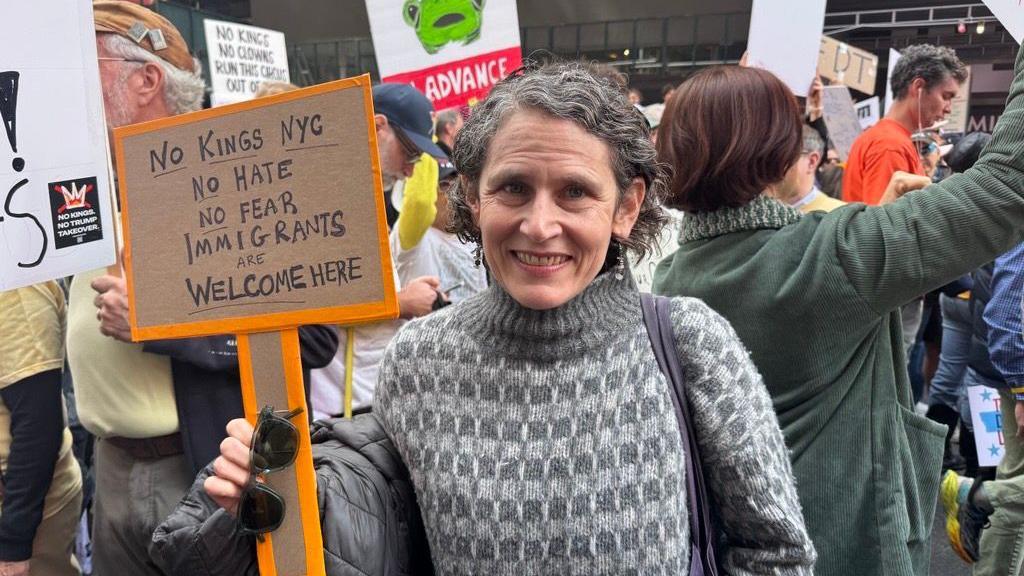 Beth smiling at camera in front of crowd of protesters, holding a sign that says "No kings NYC, no hate, no fear, immigrants are welcome here"