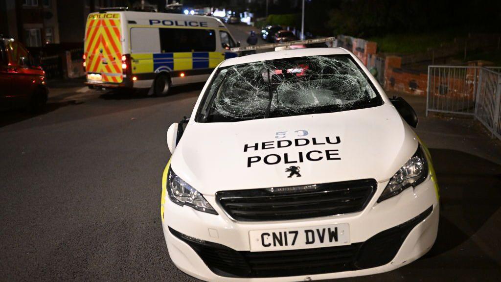 Police car with smashed front window and police van behind