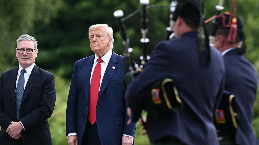 Sir Keir Starmer and President Donald Trump are standing side by side in front of two male bagpipers, who have their backs turned to us. Sir Keir is in a dark blue suit with white shirt and blue tie. President Trump is in a dark blue suit with white shirt and bright red tie. They are all standing in front of green trees.