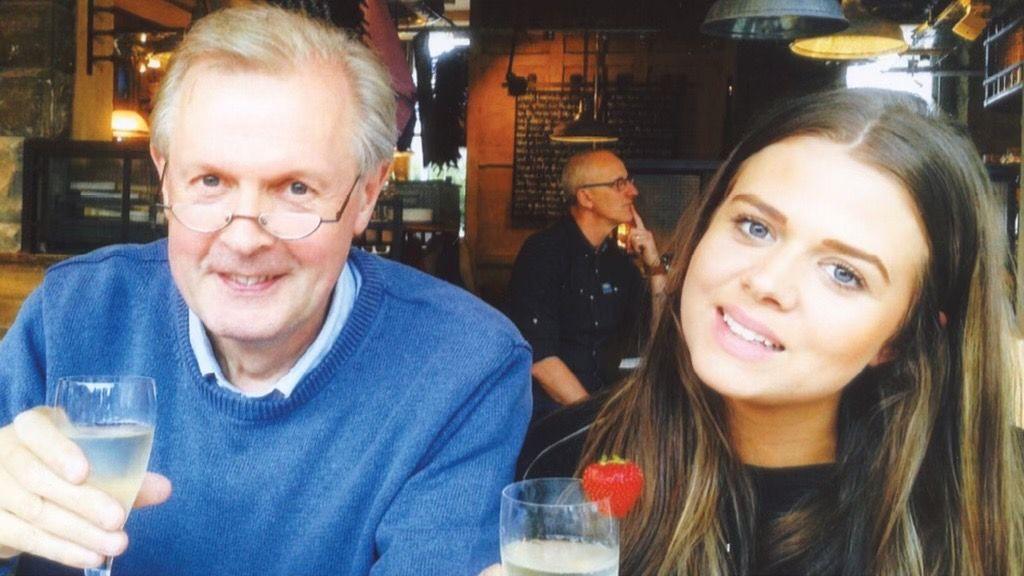 Len Noakes on the left wearing a blue jumper wearing glasses. He is holding up a glass in a restaurant smiling at the camera. He has thinning grey hair and slightly rosy cheeks. Next to him is Claire Noakes. She is smiling and has brown hair with blonde streaks running through it. She too is holding a glass and it has a strawberry hooked onto the side of it. In between them, a man is sat at a table in the background with his finger resting on his chin.