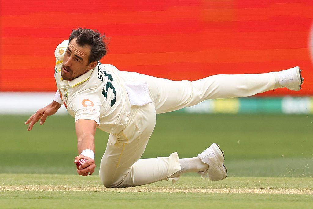 A cricketer dives forward in full stretch, clutching the ball with one hand as he lands on the pitch, showcasing athleticism and quick reflexes during a fielding effort.