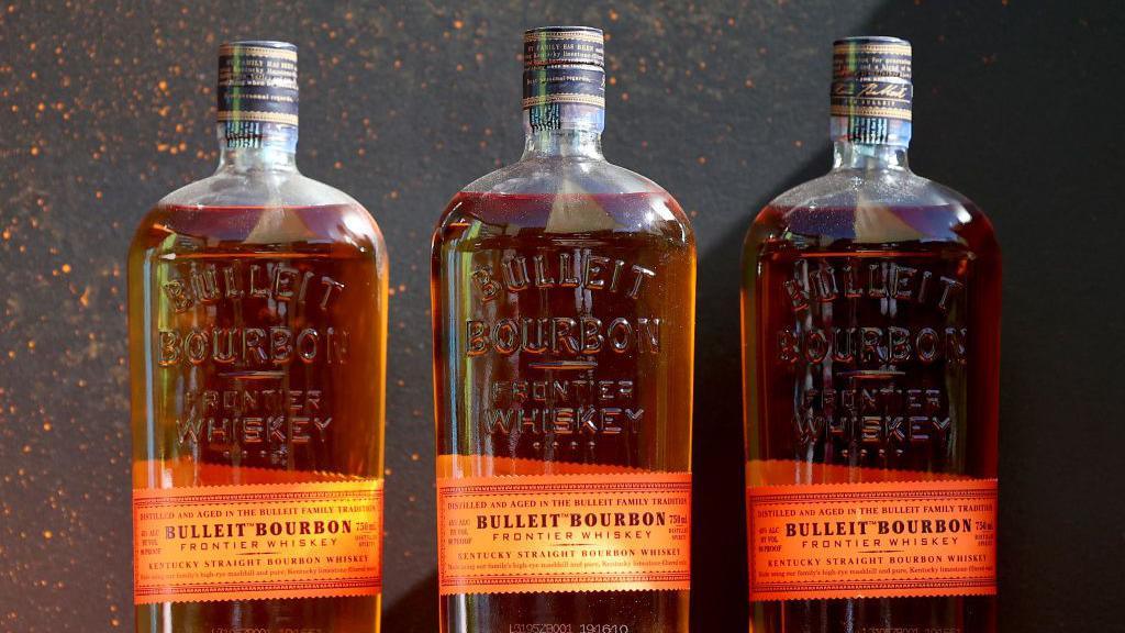 Three bottles of Bulleit Bourbon are lined up against a black stone background
The labels are orange and printed with vintage-style writing, while the bottles, filled with golden liquor, are embossed with the words "Bulleit Bourbon: Frontier Whiskey"