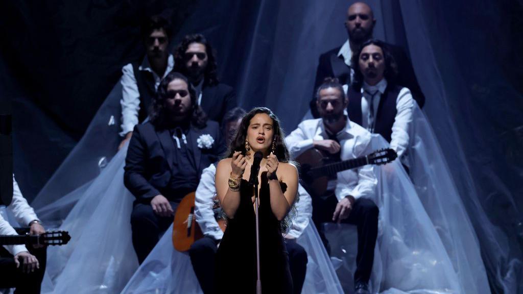 Rosalía wears a black off-the-shoulder dress. She is singing into a microphone stand while performing on stage. There are musicians with guitars sitting behind her. 
