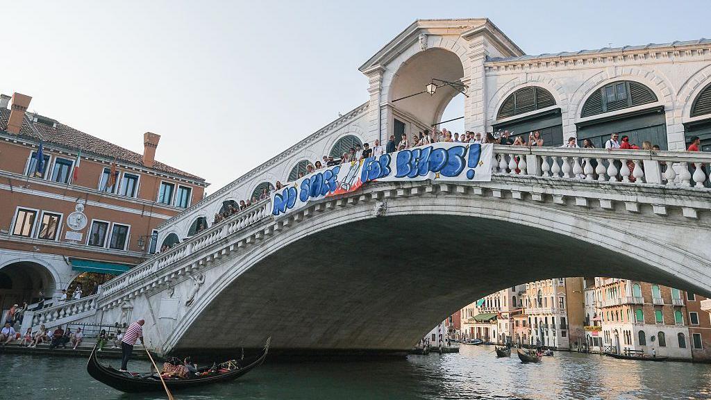 A big sign that says "no space for Bezos" is hung on a bridge in Venice that goes over a canal. There is a gondola sailing through the space under the bridge.