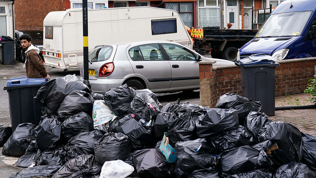 Bin bags piled up on a street in Sparkhill, Birmingham in May 2025. There is a man on the street corner, with cars, a caravan and houses in the background