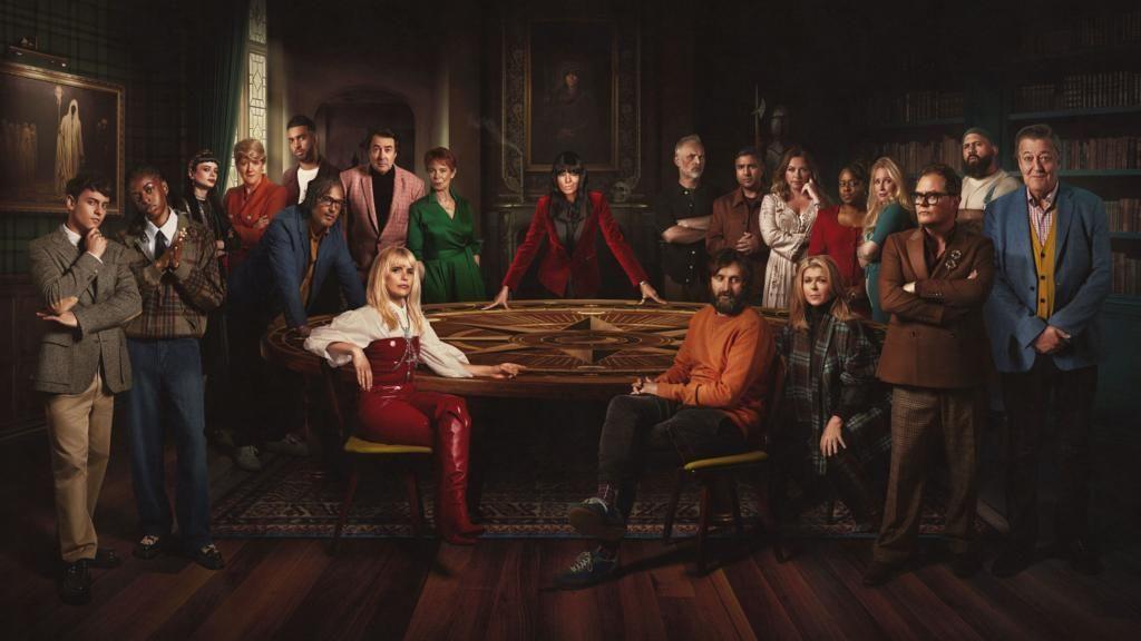 The entire cast of the Celebrity Traitors show, pictured sitting around the round table. Host Claudia Winkleman stands central, behind the table, wearing a red velvet jacket.