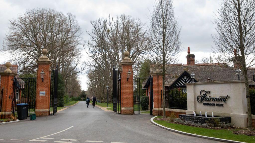 The entrance to the Fairmont Windsor Park Hotel where large pillars in red brick are holding two black metal gates and a straight road continues in to the distance. A sign to the side of the road has the name of the hotel in a script style font.