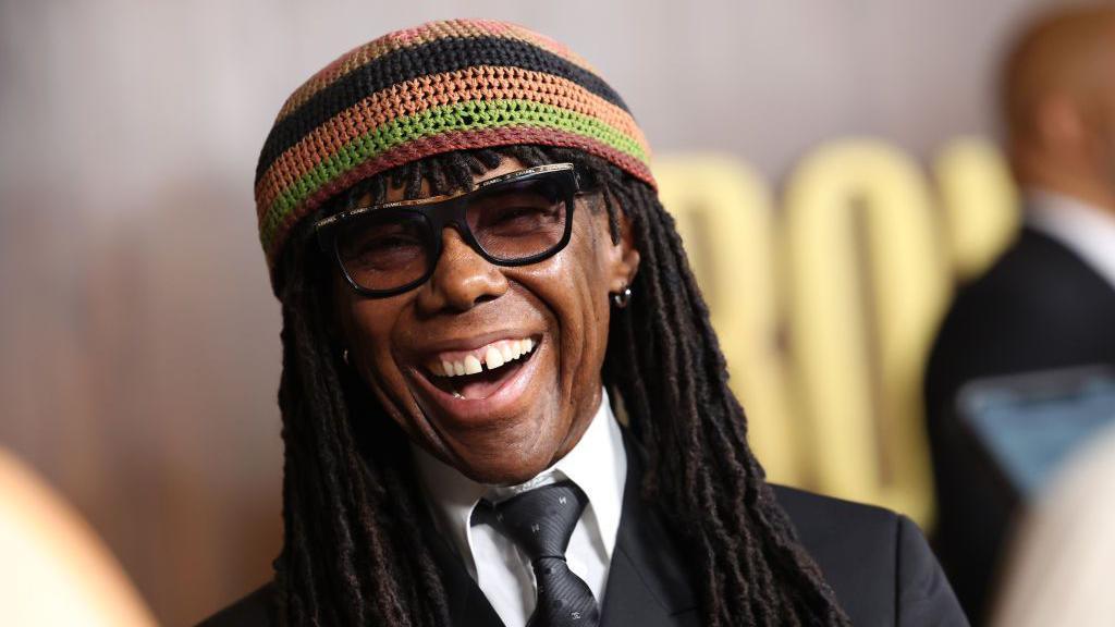 A man with sunglasses, beanie hat and black dreadlocks smiles. He is wearing a suit. 