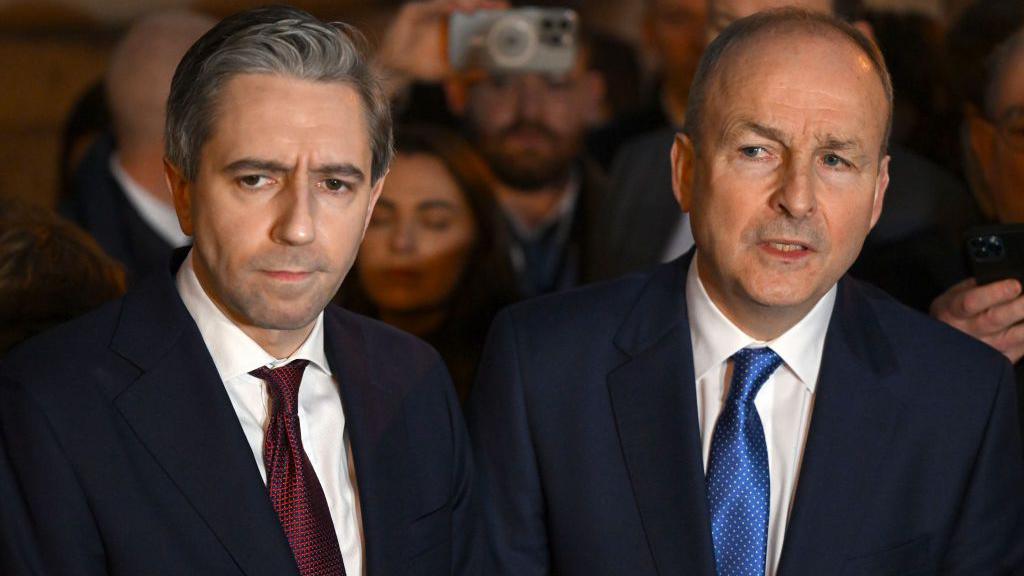 Simon Harris, who is standing beside Mr Martin, looks into the distance. Martin is balding and is wearing a dark blue suit, white shirt and royal blue tie, while Harris has short grey hair and is wearing a dark blue suit, white shirt and maroon tie.