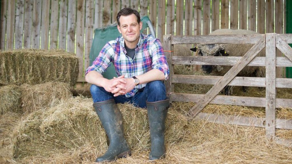 Matt has short brown hair and is wearing a pink, blue and white checked shirt, blue jeans and grey wellies. He is sitting on hay bales in a barn in front of a wooden gate. Behind him is a sheep.