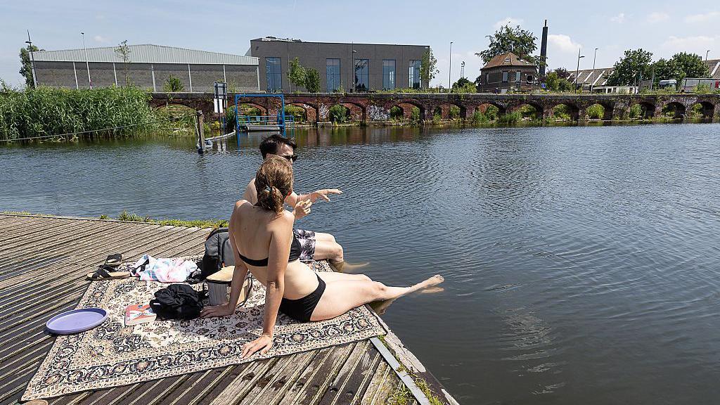 A man and woman in swimming costumes sitting on a blanket in the sunshine by a canal in Gent