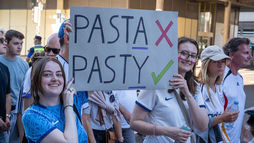 Two England fans hold up a homemade sign saying "Pasta X Pasty ✓"
