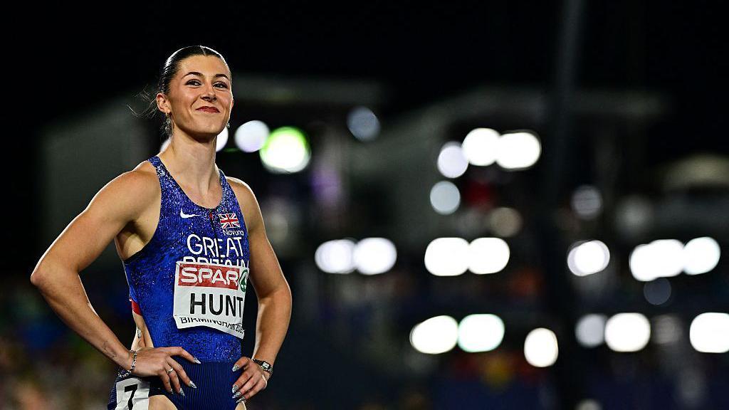 Amy Hunt smiles and looks beyond the camera with her hands on her hips. The stadium lights are blurred in the background.